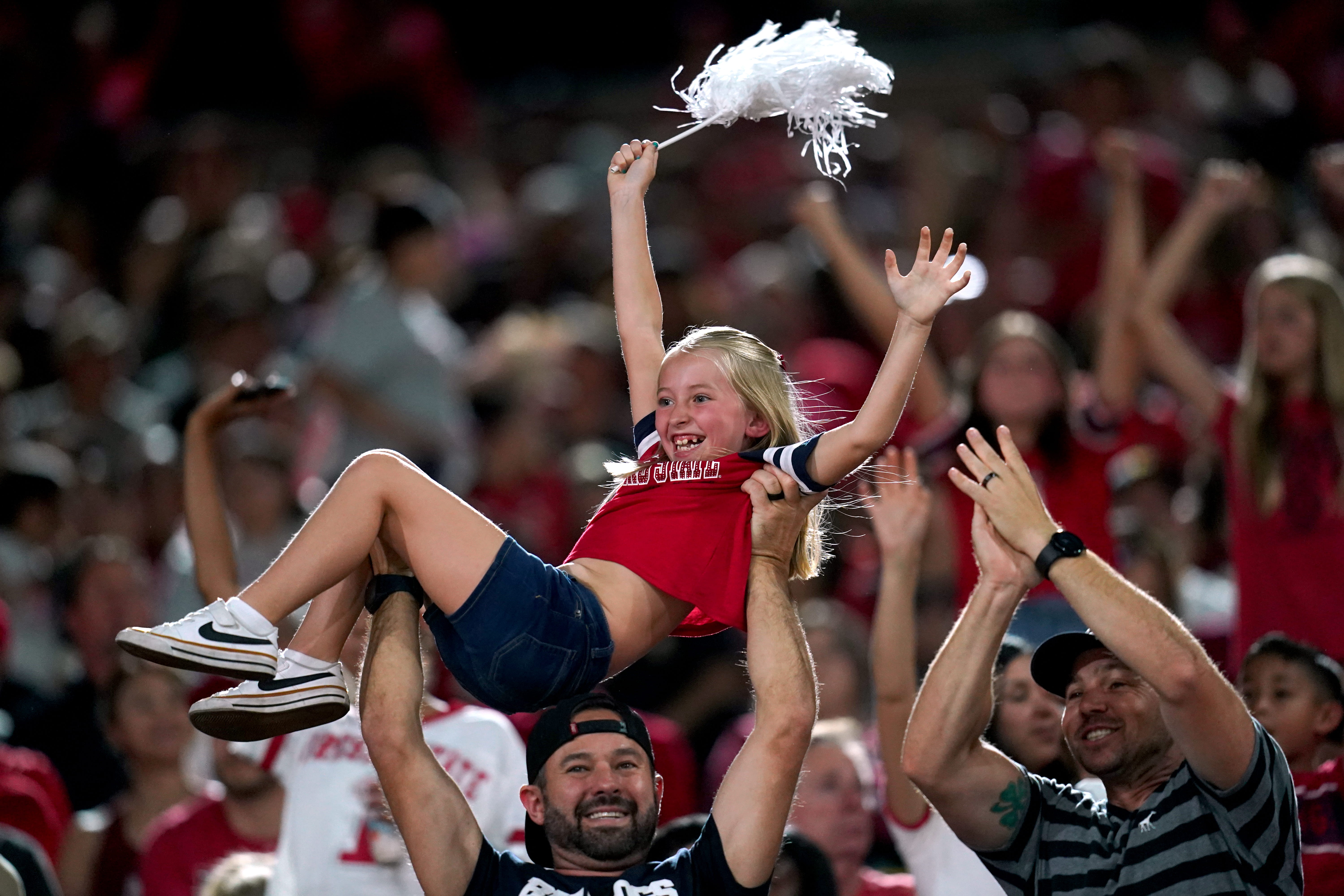 A girl lifted above a cheering crowd at a football game, holding a white pom-pom in the air with both arms raised. Fans around her celebrate, creating an energetic and spirited atmosphere.
